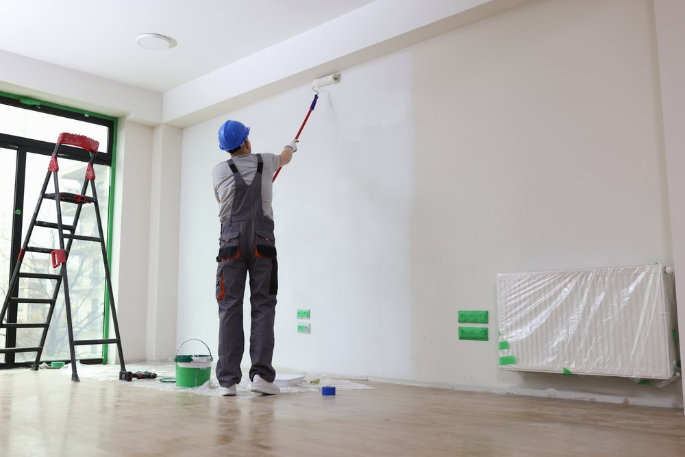 Illustratie van een persoon die renovlies aanbrengt op een muur met behulp van behanglijm, een roller en een plamuurmes.