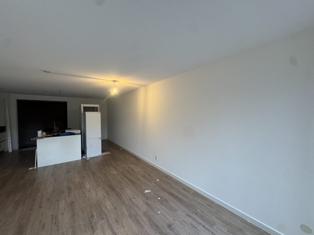 Woonkamer met gladde witte wanden, een laminaatvloer en een keukenblok op de achtergrond
