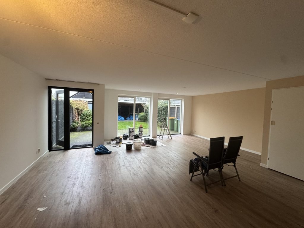 Ruime woonkamer met lichte gesausde wanden, een houten vloer en tuindeuren, met verfemmers bij de pui